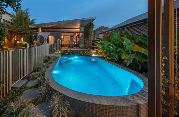Twilight view of a compact landscaped backyard featuring a curving turquoise swimming pool, dark tiled pool surround and raised wall, dense tropical planting, ornamental grasses, stepping stones, and a timber-framed outdoor entertaining pavilion with warm interior lighting. Brick boundary walls and a slatted metal fence enclose the private courtyard.