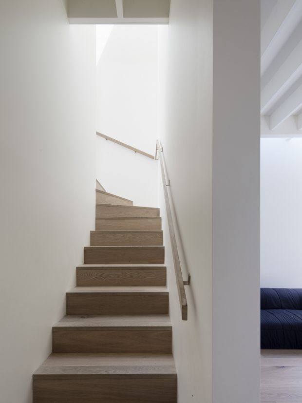 Minimalist interior photograph looking up a narrow staircase with warm wood treads, white walls, and simple timber handrails. Bright natural light enters from above, while part of an adjacent room with a dark blue sofa and pale timber flooring is visible through the opening on the right.