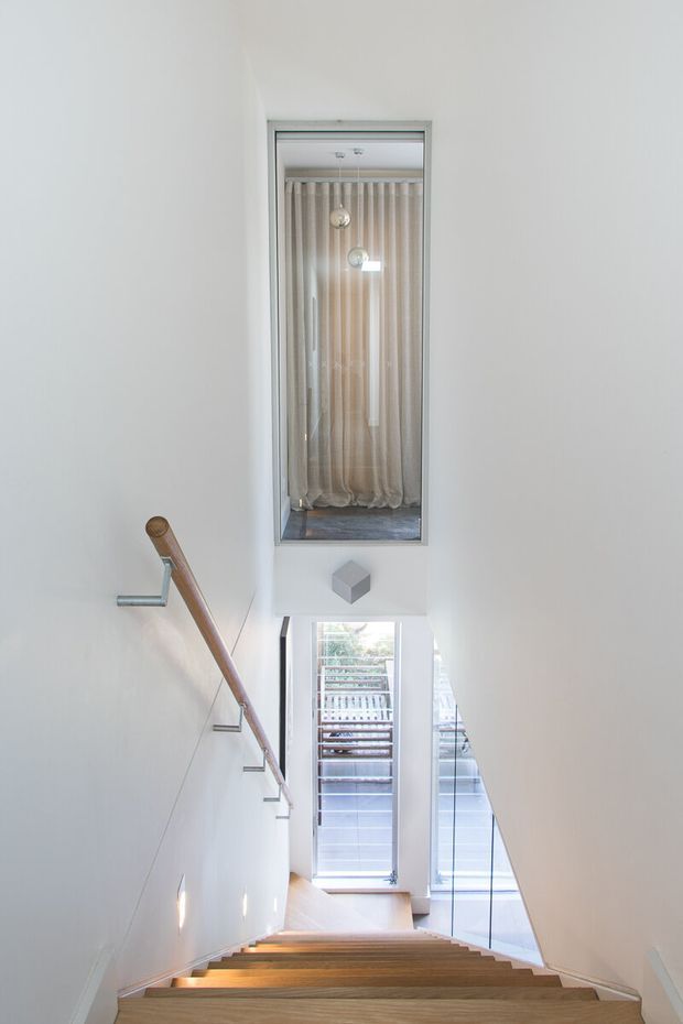 A bright, minimalist stairway viewed from above, with warm timber treads, white walls, a simple timber handrail, and frameless glass balustrading. Tall glazing and an upper internal window with sheer curtains bring in abundant natural light, creating a clean, airy vertical circulation space.