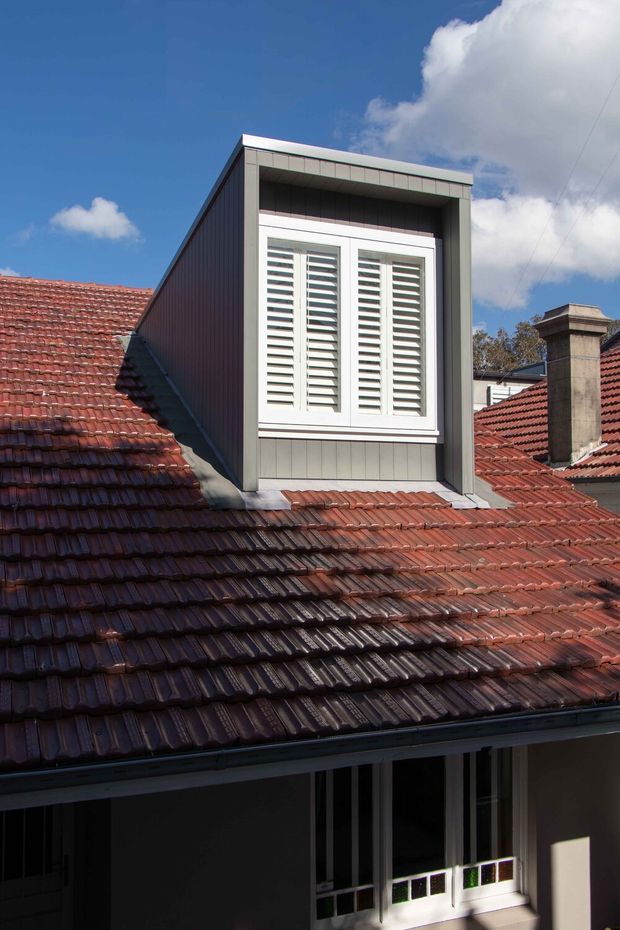 Exterior architectural photograph of a residential roof addition or dormer. A steep red terracotta tiled roof is dominated by a contemporary box-like dormer clad in dark grey vertical panels, with a white louvred window and deep projecting frame. Older tiled roofs, a masonry chimney, and part of the lower facade are visible beneath a bright blue sky.