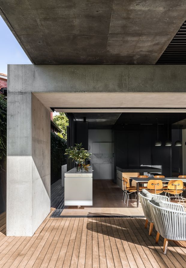 A contemporary indoor-outdoor dining and kitchen space opening onto a timber deck. The structure features a large exposed concrete overhang and walls, dark cabinetry, a marble-look splashback, timber dining furniture, pendant lighting, and a partially enclosed outdoor lounge area. Strong contrast between the sunlit deck and the moody interior.