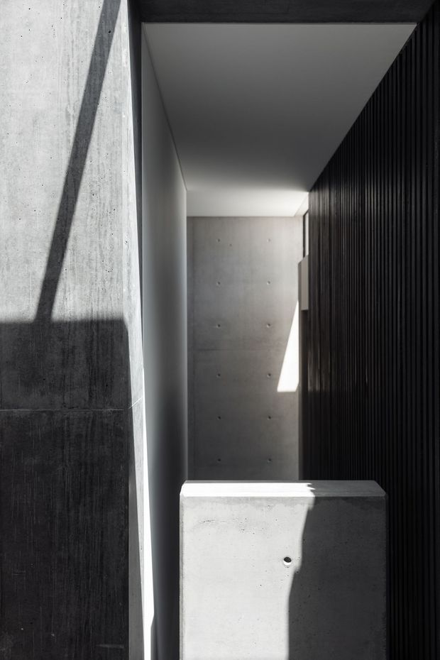 A narrow, dramatic architectural interior or passageway composed of raw concrete walls and a freestanding concrete block or bench in the foreground. A tall black vertical slatted wall lines the right side, while strong sunlight creates sharply defined geometric shadows across the concrete surfaces. The restrained monochromatic palette and minimal detailing create a sculptural, highly atmospheric composition.