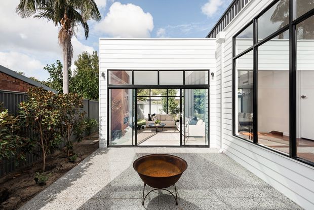 Contemporary white weatherboard-clad residential extension arranged around a gravel courtyard. Large black-framed glazed doors and windows connect the courtyard to a bright living area, while a sculptural fire bowl provides a focal point in the foreground. Palm planting and neighbouring brickwork reinforce the warm outdoor setting.