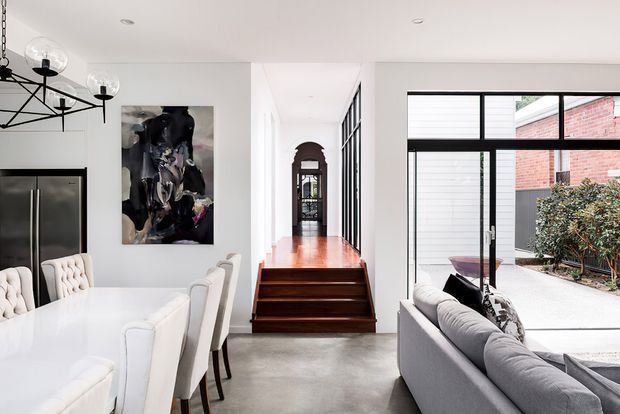 Bright open-plan interior showing a dining area and living room arranged around a central hallway. The space features a white dining table with upholstered chairs, a grey sofa, polished concrete flooring, white walls, black-framed glazing and doors, stainless-steel kitchen appliances, a contemporary suspended chandelier, and timber steps leading to an arched corridor. A courtyard and adjacent brick wall are visible through the glazing.