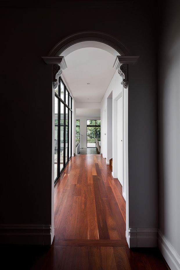 A long, elegant interior hallway viewed through a dark, ornate arched opening. The corridor has polished reddish-brown timber flooring, white walls and ceiling, black-framed floor-to-ceiling glazing along the left side, and white paneled door openings on the right. The passage leads toward a bright living area with garden views, combining traditional decorative arch detailing with contemporary glazing and finishes.