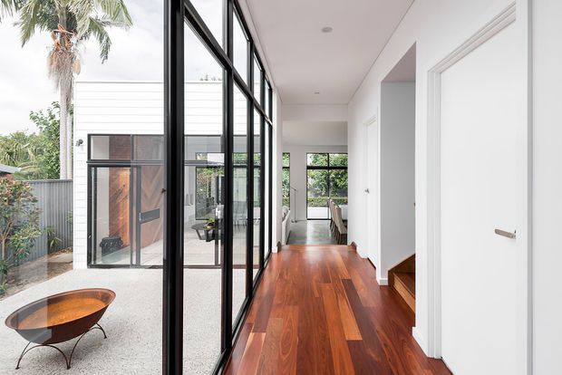 A bright modern residential hallway with rich timber flooring, white walls and ceiling, and extensive black-framed floor-to-ceiling glazing along one side. The glazing opens views to a landscaped courtyard with a sculptural outdoor fire bowl and palm tree. A dining area is visible at the far end, and a short set of timber stairs leads upward beside white interior doors.