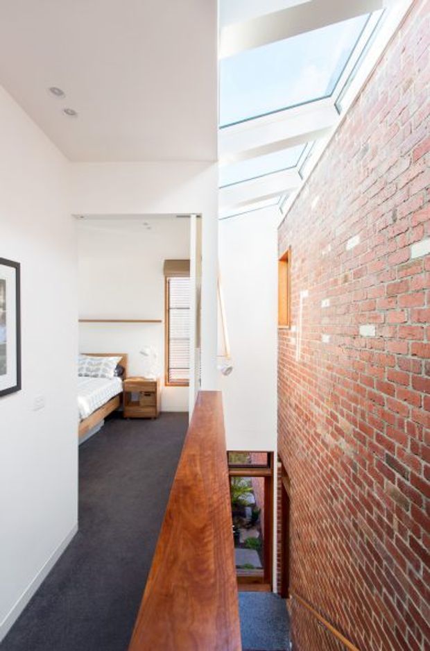 A bright, narrow interior landing or hallway overlooking a lower level, with a long polished timber balustrade beside an exposed red-brick wall. A series of roof skylights brings in strong daylight. Through the opening, a bedroom with a bed, dark carpet, timber bedside table and window blind is visible. The space has a contemporary renovation character with retained masonry and warm timber detailing.