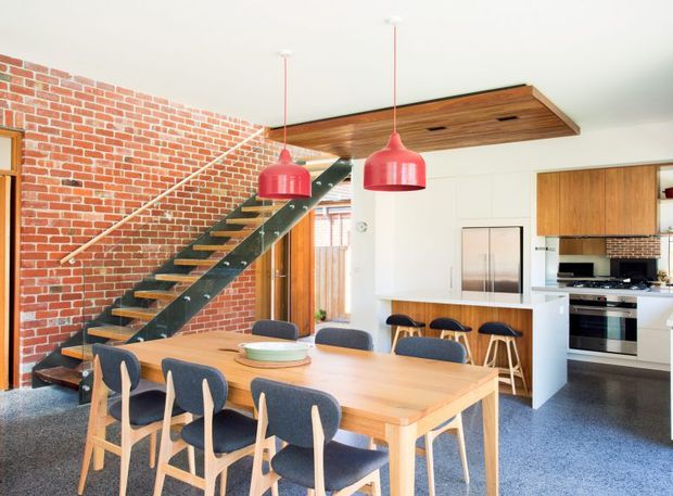 Bright open-plan dining and kitchen interior with a timber dining table and upholstered timber chairs in the foreground. A contemporary kitchen with white cabinetry, timber joinery and stainless-steel appliances sits behind the dining area. A striking exposed red-brick feature wall supports an open-riser staircase with dark metal stringers and timber treads. Timber ceiling panelling and red pendant lights add warmth and colour.