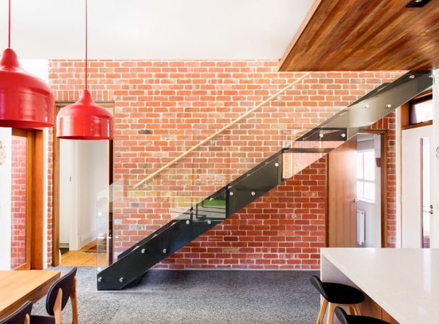 A bright industrial-style interior centered on a black steel open-riser staircase with a clear glass balustrade, set against an exposed red brick wall. Warm timber ceiling lining and joinery contrast with the brick and dark stair structure. Red pendant lights hang over a timber dining area at left, while a white workbench or kitchen counter with stools occupies the right foreground. A doorway and interior glazing are visible beneath and beside the stairs.