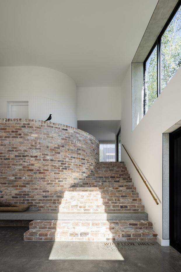 A bright contemporary interior focused on a sweeping curved brick stair and adjoining curved brick half-wall. Broad brick steps rise toward a corridor beneath a high white ceiling, with a simple brass-toned handrail, dark concrete floor, exposed concrete window reveals, and tall black-framed glazing bringing in strong sunlight. The space has sculptural geometry and a restrained material palette of pale plaster, reclaimed-looking brick, concrete, and dark metal.