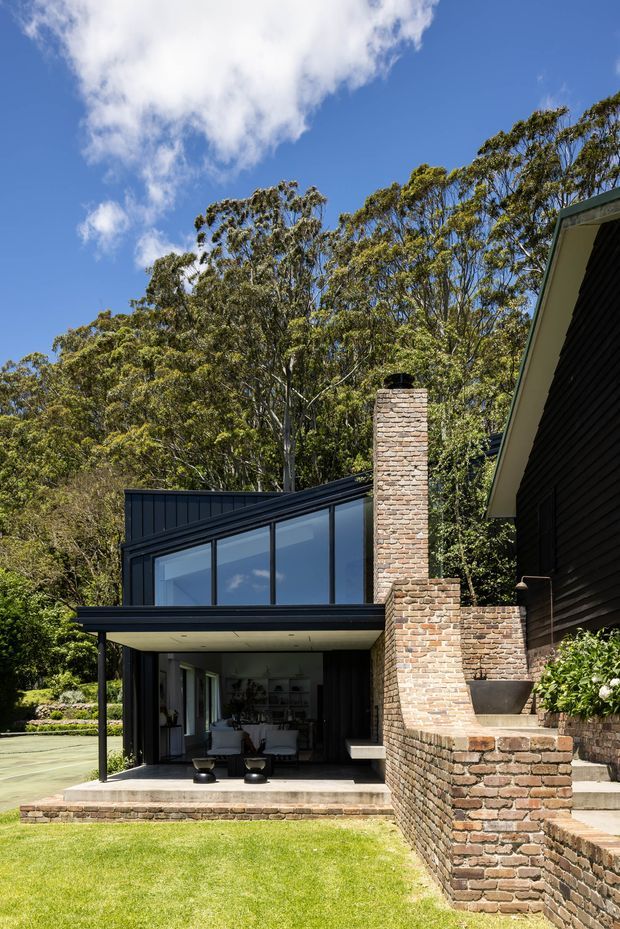 Exterior view of a contemporary dark-clad house set within a leafy landscaped site. The building features a steeply pitched roof, extensive floor-to-ceiling glazing, a projecting covered terrace, dark vertical cladding, and a prominent exposed brick chimney with integrated brick garden or retaining walls. A furnished living area is visible through the glazing, with lawn and mature trees surrounding the home.