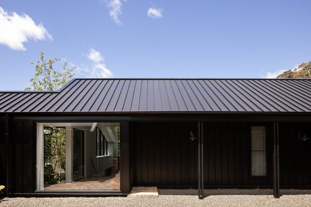 Exterior view of a compact, low-profile building with a dark vertically clad facade and a pitched standing-seam metal roof. Large glazed sliding doors open toward a light-toned interior, while dark-framed windows and simple exterior wall lights reinforce the restrained contemporary design. The building sits beside a pale gravel surface with trees visible around it under a clear sky.