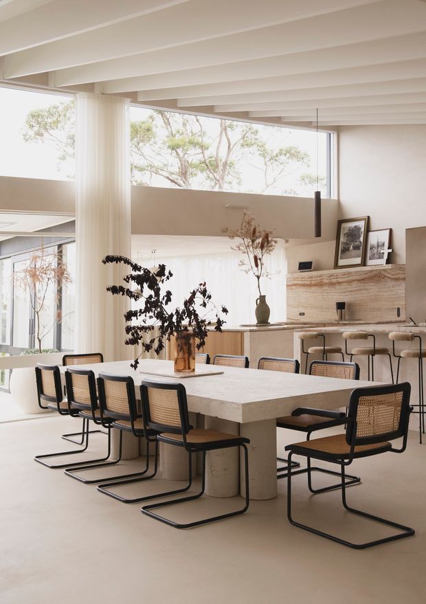 A bright, spacious open-plan dining and kitchen interior with a large pale stone dining table surrounded by black-framed cane dining chairs. A warm neutral kitchen with a long island and bar stools sits behind it, complemented by a textured stone fireplace or feature wall, framed artwork, sculptural pendant lighting, extensive glazing, and exposed painted ceiling beams.