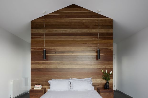 A contemporary bedroom viewed straight on, featuring a bed against a dramatic full-height horizontal timber feature wall shaped to follow the pitched roofline. Two slim black pendant lights hang beside the bed, with timber bedside tables, white bedding, white walls, and dark flooring. A small vase of greenery adds a natural accent.
