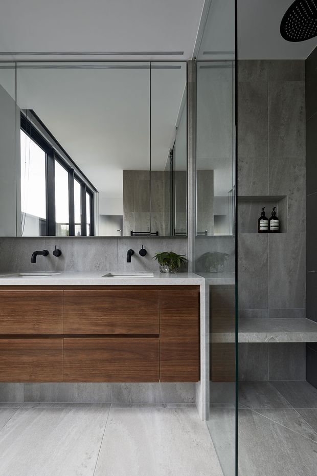 A contemporary bathroom with a floating double vanity in warm timber cabinetry, a pale stone benchtop, and black wall-mounted tapware. Large mirrored panels reflect the room and black-framed glazing. To the right is a frameless glass shower enclosure clad in large grey stone-look tiles, with a recessed shelf and built-in bench. The palette is restrained and refined, combining grey surfaces, timber joinery, black fixtures, and clear glass.