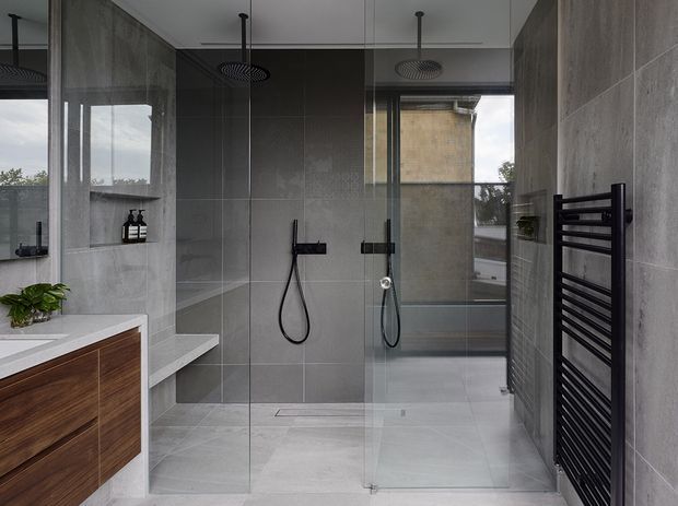 Contemporary grey-tiled bathroom with a frameless glass walk-in shower, dual overhead rain showers, wall-mounted hand showers, floating timber vanity with a white benchtop, recessed niche shelving, and a black heated towel rail. Natural light enters through large windows, creating a refined, cool-toned space.