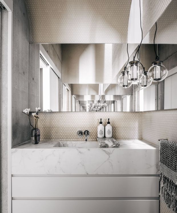 Contemporary bathroom vanity with a white marble-look benchtop, integrated basin, pale hexagonal mosaic tile splashback and matching tiled ceiling detail. A large mirror creates a repeated corridor-like reflection, while globe pendant lights hang above the vanity. Concrete-look wall surfaces, minimalist white drawers, chrome tapware and monochrome accessories create a refined, modern atmosphere.