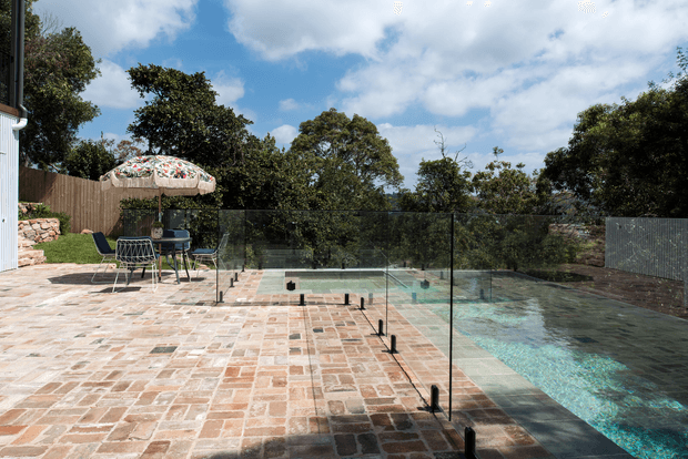 A landscaped outdoor pool area with a long rectangular swimming pool bordered by frameless glass safety fencing and a broad, warm-toned paved terrace. A small outdoor dining setting with a patterned umbrella sits beside the pool, with mature trees and a timber privacy fence beyond.
