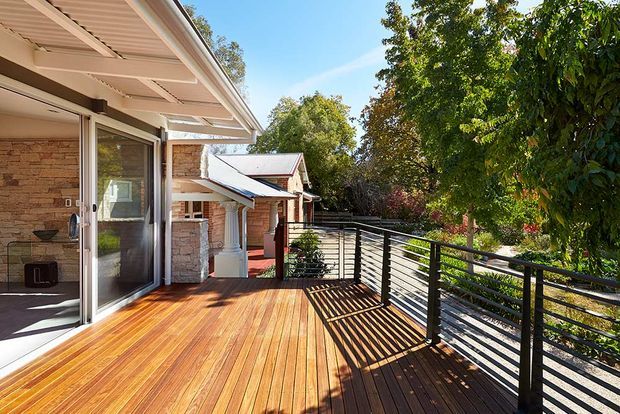 Sunlit timber deck beside a brick-clad house, with open glazed doors leading indoors and a black horizontal metal balustrade overlooking a leafy garden. The deck has a covered roof extension and connects to a small secondary roofed structure, creating an inviting outdoor entertaining area.