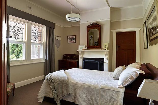 A traditional bedroom with a neatly made bed, white textured bedding, dark wood bed frame, and upholstered headboard. The room features a decorative white fireplace mantel with a dark inset, an ornate overmantel mirror, timber doors, framed artwork, decorative leadlight-style windows, heavy curtains, and classic ceiling detailing. A modern circular pendant light provides contrast with the heritage-inspired interior.