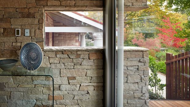 Interior view featuring a rough stacked-stone wall, a slim glass console table, and a decorative patterned plate. A large glazed opening looks onto a matching stone exterior wall, timber decking, a dark timber fence, and landscaped planting. The image has a natural, earthy material palette and strong indoor-outdoor connection.