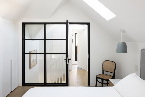 Bright, minimalist bedroom viewed toward a black-framed glazed internal partition and open doorway leading to a hallway or stair area. The room has a sloping white ceiling with a large skylight, pale timber flooring, a white bed, a woven-back black chair, and a simple pendant light. Strong natural light and restrained detailing create an airy contemporary interior.