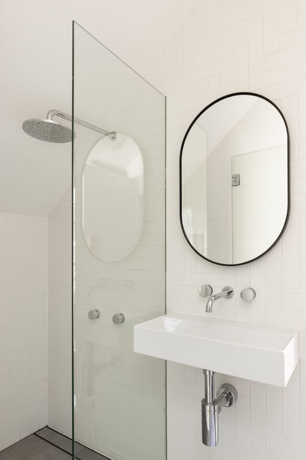 Bright, compact contemporary bathroom featuring a frameless glass walk-in shower, white subway-tile walls, a wall-mounted rectangular white basin with exposed chrome plumbing, and a large black-framed rounded-arch mirror. The palette is predominantly white with polished chrome and subtle grey flooring.