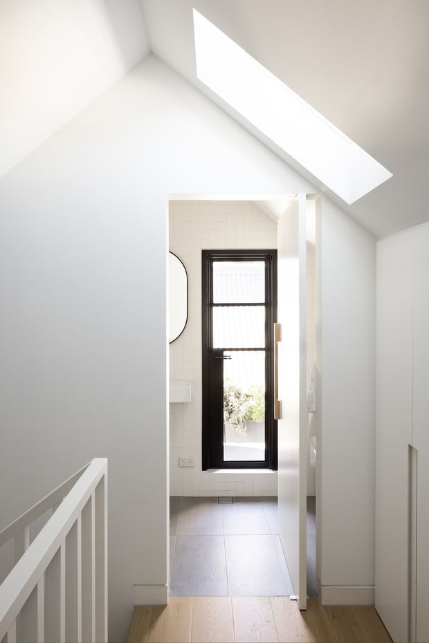 Bright minimalist hallway or landing viewed from a stairway, with white walls, a white balustrade, pale timber flooring, built-in white cabinetry, and a glazed black-framed door at the end. A large angular skylight brings strong natural light into the space. The doorway opens toward a small tiled room or entry area with white wall tiles and a narrow vertical window.