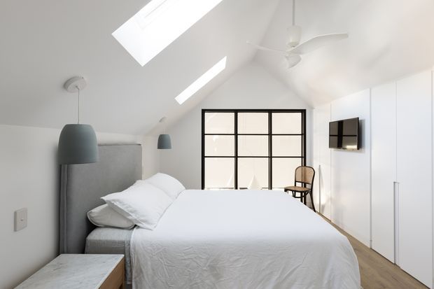 Bright, minimalist bedroom with a pitched white ceiling and two skylights, a large upholstered grey bedhead, white bedding, pale timber flooring, and full-height white built-in wardrobes. Black-framed internal glazing forms a partition at the far end, with a small timber chair, wall-mounted television, pendant lights, and ceiling fan.