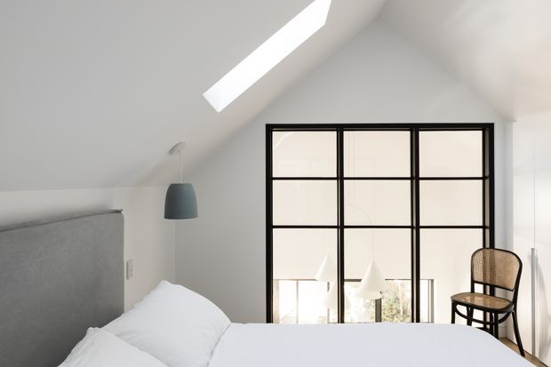 Bright minimalist bedroom beneath a sloped ceiling, featuring a partially visible upholstered grey headboard, white bedding, a dark pendant light, a roof skylight, and a black-framed glazed partition with a woven chair beyond.