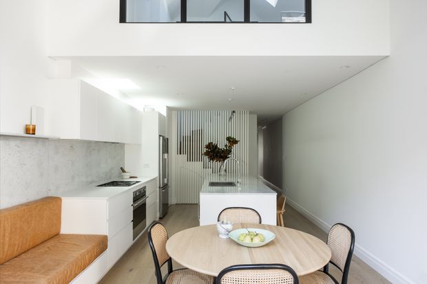 Bright contemporary open-plan kitchen and dining area with white cabinetry, a pale concrete-look splashback, integrated appliances, a central island, and a round timber dining table with woven chairs. A warm tan built-in bench seat sits along the left wall. Vertical slatted screening and a high-level internal window add architectural detail, while a hallway extends beyond the kitchen.