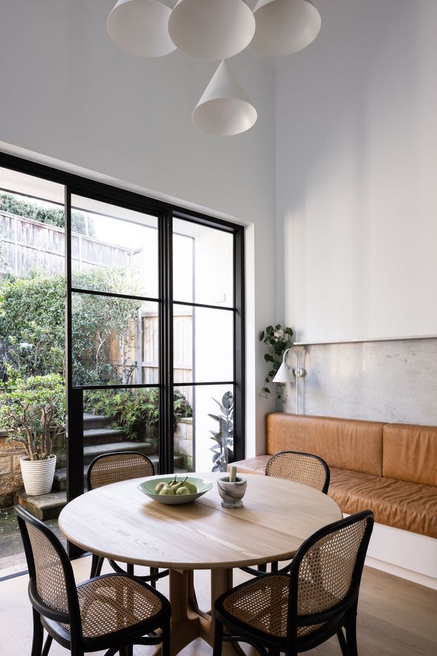 Bright, refined dining nook with a round light-wood dining table surrounded by black-framed cane-back chairs. A built-in tan leather banquette sits against a pale concrete-look wall, beneath a sculptural white pendant light. Full-height black-framed glazing opens toward a planted courtyard, while potted greenery and soft neutral finishes create a contemporary, warm atmosphere.
