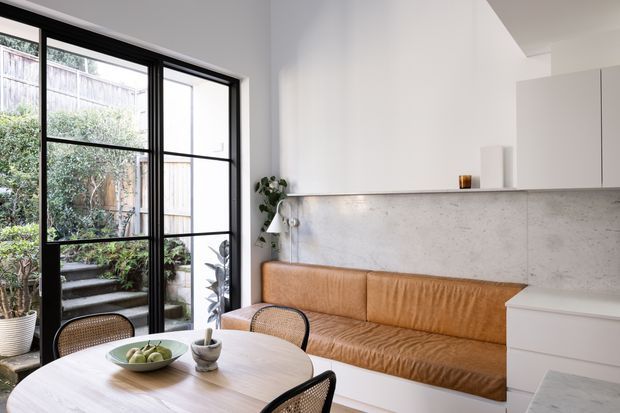 A bright, compact contemporary dining and living space with a pale timber dining table, woven-back chairs, a built-in tan leather banquette, and minimalist white cabinetry. Large black-framed glazed doors open to a planted outdoor stairway, while a pale textured stone or concrete backsplash forms a feature wall behind the seating. The room has a clean, warm and natural aesthetic with strong indoor–outdoor connection.