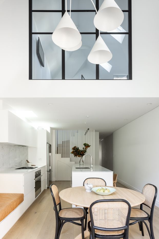 Bright, minimalist open-plan kitchen and dining area with white cabinetry, pale timber flooring, a white island, and a round light-wood dining table surrounded by woven cane chairs. Large black-framed internal glazing reveals a stairwell or adjacent upper-level space, while sculptural white pendant lights hang above. A vertical slatted screen and floral arrangement add visual interest.