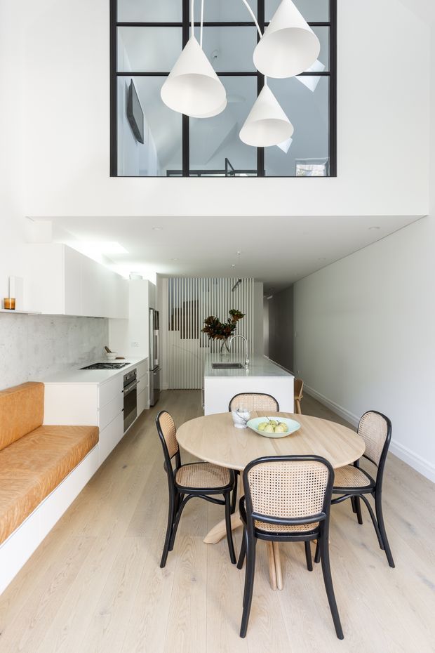 Bright open-plan kitchen and dining area with white cabinetry, pale timber flooring, a light stone island and backsplash, and a round timber dining table surrounded by black-framed cane chairs. A built-in caramel upholstered bench lines the left wall. Three sculptural white pendant lights hang beneath a high internal glazed opening with black mullions, creating a clean, airy contemporary interior.