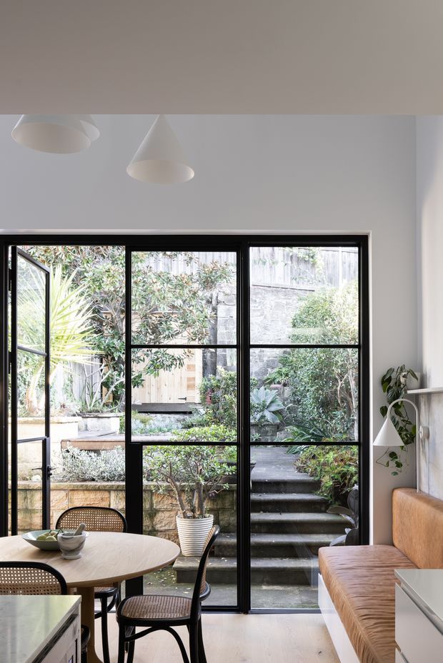 Bright, compact dining area beside full-height black-framed glazing and a hinged glass door opening onto a landscaped courtyard with terraced steps. The room includes a round light-wood dining table with woven cane chairs, a built-in caramel upholstered bench, white cabinetry, pale flooring, simple pendant lights, and a small wall-mounted lamp with trailing plant. The interior has a clean contemporary character with strong indoor-outdoor connection.