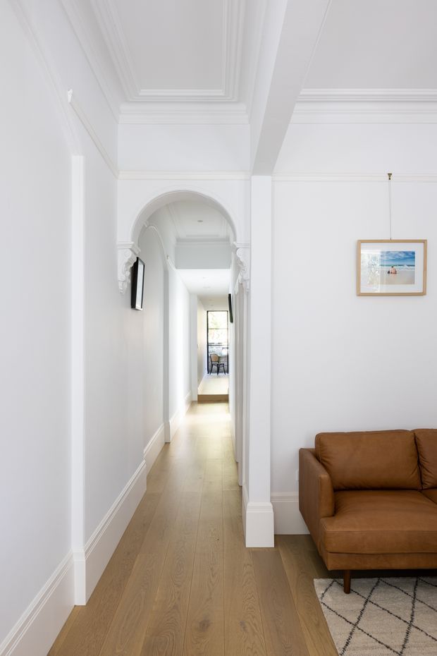 Bright, refined residential hallway viewed from an adjoining living area. The corridor has pale timber flooring, white walls, ornate ceiling cornices and panelling, a decorative arched opening with carved trim, and a framed artwork. Part of a caramel-brown sofa and patterned rug are visible in the foreground, with daylight drawing the eye toward a distant seating area.