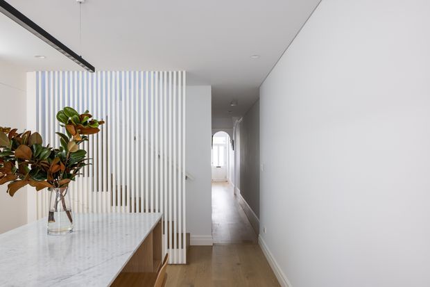 Bright contemporary interior showing a dining area opening onto a long hallway. A white marble dining table with timber joinery sits in the foreground, beside a tall indoor plant and a vertical white slatted screen that partially separates the spaces. The hallway has light timber flooring, white walls and ceiling, recessed lighting, and an arched opening visible at the far end.