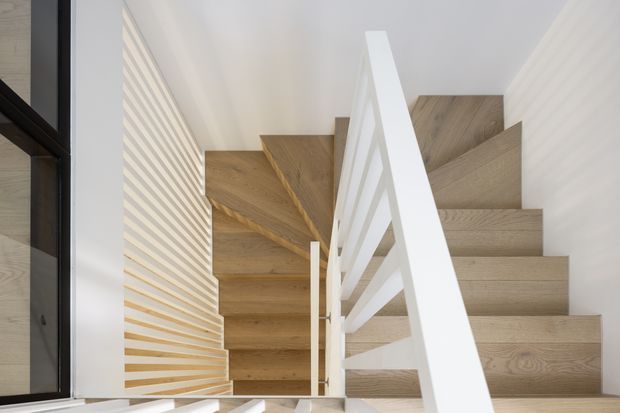 A bright modern stairway viewed from above, featuring pale timber treads and risers, a crisp white open balustrade, and a textured timber slat feature wall along the stairwell. Black-framed glazing is visible at the left edge. The clean lines and light neutral palette create an airy, contemporary interior.