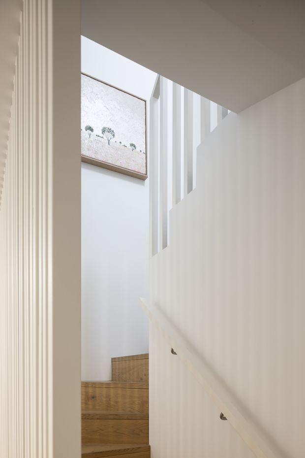 Bright, minimalist stairway viewed from a hallway or adjoining space. The staircase features warm natural timber treads, white painted walls, a solid white balustrade with vertical openings, and a simple timber-framed landscape artwork on the upper wall. Soft daylight enters through the stairwell, creating an airy and calm atmosphere.