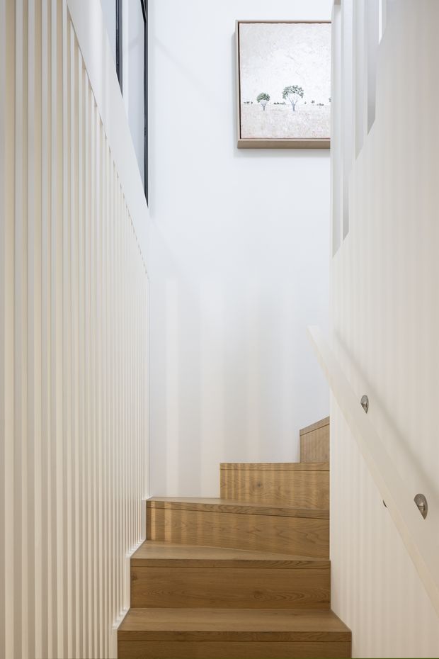 A bright, narrow interior stairway with warm timber treads and risers, white walls, and vertical white slatted wall panelling. A white handrail runs up the right side, while a framed minimalist landscape artwork is mounted above the stairs. The space is clean, light-filled, and restrained in design.