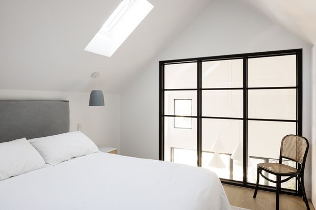 Bright, minimalist bedroom with a sloped white ceiling, skylight, grey upholstered bedhead, white bedding, and a large black-framed glazed partition or window. A woven-seat chair and simple pendant light add restrained contrast, while daylight creates an airy atmosphere.