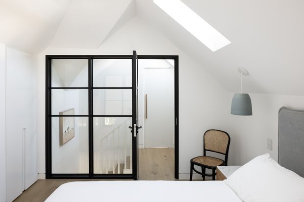 Bright, minimalist bedroom beneath a sloped ceiling, featuring a skylight, white walls, pale timber flooring, and a grey upholstered bed. A black-framed glazed internal door and partition open toward a stair landing, while a woven-seat accent chair and pendant light add restrained contrast.