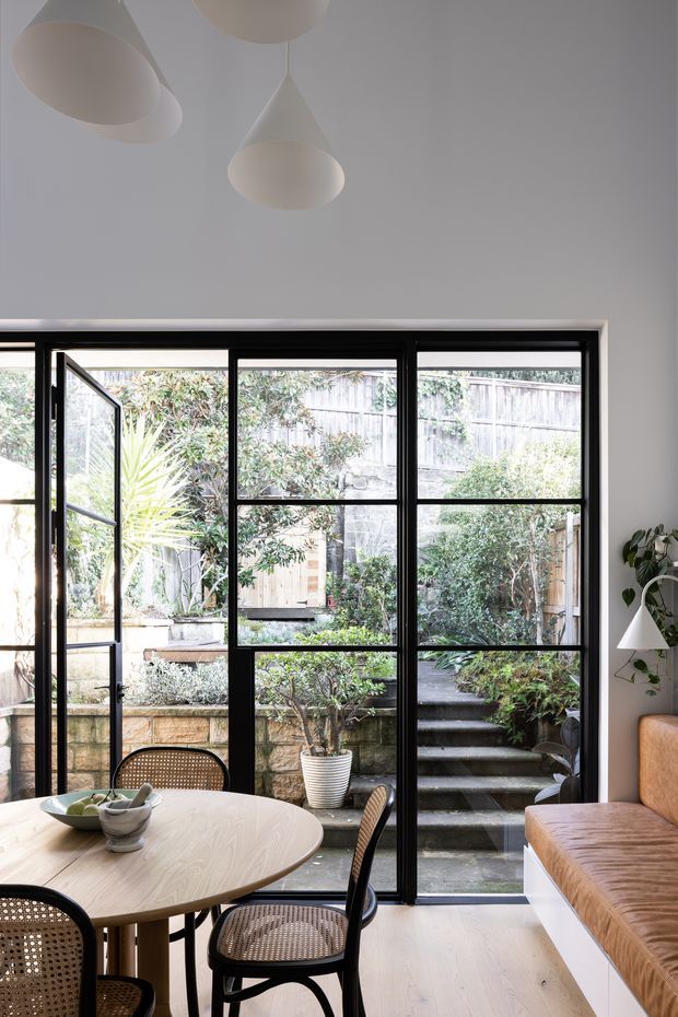 A bright, compact dining and living area with a pale timber oval dining table, black cane dining chairs, and a tan upholstered built-in bench seat. Large black-framed glazed doors and windows open onto a planted courtyard with steps and timber fencing. The room has white walls and ceiling, light timber flooring, sculptural white pendant lights, and a small wall-mounted lamp with a potted plant. The overall look is calm, contemporary, and softly minimalist.