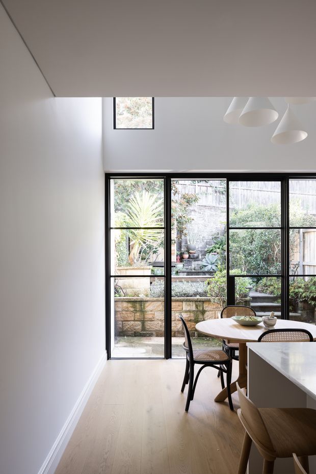 A bright, narrow contemporary dining area beside a kitchen, with pale timber flooring, white walls and ceiling, black-framed full-height glazed doors and windows opening to a planted courtyard. A round light-wood dining table with woven black chairs sits near the glazing, while a pale stone or composite kitchen island occupies the foreground. Minimal white pendant lights hang above, and a small high-level window adds natural light.