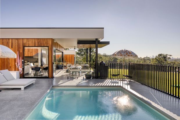 A contemporary elevated outdoor terrace with a compact rectangular swimming pool in the foreground, connected to a timber-clad house through large glazed sliding doors. The terrace includes outdoor seating and a covered dining area, while a dark vertical-slatted balustrade frames views toward landscaped surroundings and a distinctive domed structure in the distance. Bright daylight, clean lines, and a restrained natural-material palette create a relaxed modern resort-like setting.