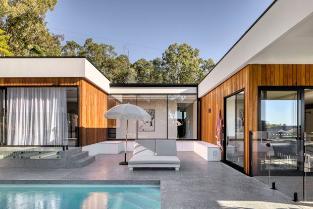 A contemporary single-storey courtyard residence arranged around a swimming pool. The exterior combines warm vertical timber wall cladding, crisp white rendered volumes and extensive dark-framed glazing. A grey paved terrace surrounds the pool and includes a sun lounger and outdoor dining area, with mature trees visible beyond the property.