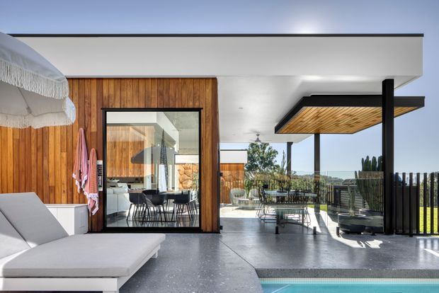 A bright contemporary outdoor entertaining area beside a swimming pool, with a covered terrace, dining setting, timber-lined ceiling, black structural posts and balustrades, and seamless glazing into a timber-clad interior. The space overlooks a landscaped suburban setting and uses a restrained palette of white, natural timber, charcoal and grey concrete.