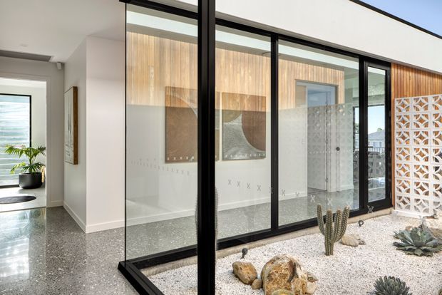 Interior view of a contemporary home featuring a glazed courtyard or lightwell enclosed by black-framed floor-to-ceiling glass panels. A white decorative breeze-block screen, white gravel, rocks, and sculptural succulents form the courtyard, while warm vertical timber wall cladding and patterned glazing are visible beyond. Polished grey terrazzo flooring continues through the adjacent hallway and living area, creating a bright, refined indoor-outdoor composition.