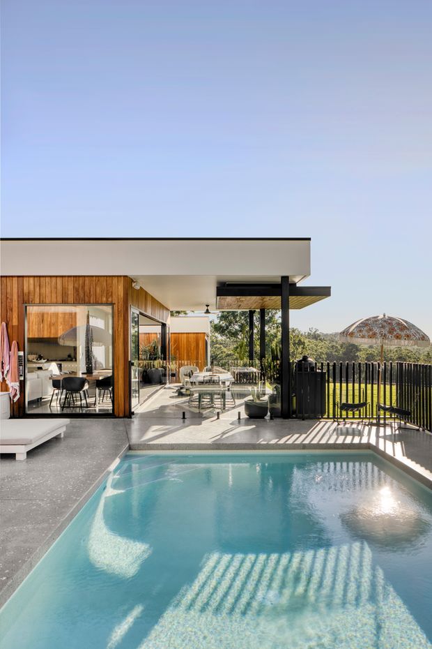 A bright contemporary residential outdoor entertaining area with a swimming pool in the foreground, a covered terrace and outdoor dining zone beside the house, timber-clad walls, extensive glazing, black structural posts and balustrades, and a flat roof. The setting includes a manicured lawn and a distinctive domed structure in the background.
